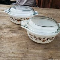 Two white Pyrex oval casserole dishes with clear glass lids on wooden surface, decorated with brown floral and butterfly pattern