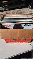 Box of vinyl records standing upright inside a cardboard box, showing partially visible record titles and worn edges.