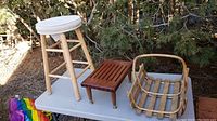 Photo showing tall wooden stool with white cushioned round seat, small wooden slatted bench, and slatted wooden basket placed on table outdoors.