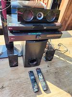 Photo of the LG 3D Blu-ray player stacked with center channel speaker on top, subwoofer in front, and multiple satellite speakers arranged around on wood surface. Two remotes placed in front.