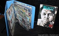 Blue baseball collector's card album opened showing several plastic sleeves full of vintage baseball cards, handheld for clear view, with Beckett price guide magazine beside it
