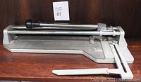Manual tile cutter with long metal cutting arm and black handle, metal base and measuring scale visible.