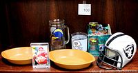 Photo showing vintage football serving bowls, Chargers glass beer stein, Raiders glass, Joe Montana football trading card in plastic stand, Raiders collectible helmet, and box of assorted football trading cards.