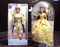 Two Disney dolls in original packaging side-by-side: Belle in yellow dress and Aladdin in satin outfit.