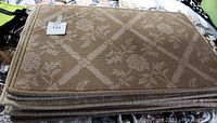 Stack of carpet door mats showing top mat with floral pattern, beige color, sewn edges.