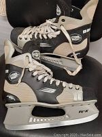Pair of Bauer Vapor ice skates in black, gray, and white colors showing the full side angle of both skates placed on a black chair, with visible Tuuk blade holders and steel blades.