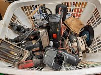 Photo showing a white plastic basket filled with a variety of used power tools including drills and a jigsaw. Some tools have visible brand labels and cords are tangled.