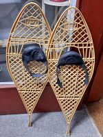 Pair of plastic snowshoes standing upright against a door, light tan in color with black adjustable foot straps.