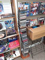 Tall curved wooden stand densely packed with around 80 CDs in jewel cases stacked vertically. Surrounding shelves have books and magazines. Box nearby with additional items.