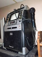 Front angled view of karaoke machine showing controls, screen, cassette player, and attached microphone.
