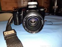 Front view of black Minolta Maxxum 5000i camera with zoom lens and Classic branded strap laid out in front.