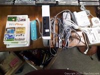 Top view of Nintendo Wii console, one blue Wii Remote, and stack of Wii game cases on a table with cables