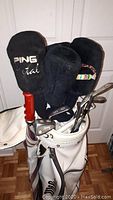 Golf clubs standing inside a white Wilson golf bag with various headcovers, including Ping, and metal shafts visible