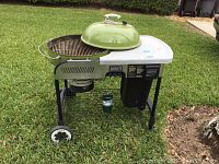 Weber green lid gas grill with attached side table and wheels on grass outdoors. Small green propane bottle mounted below grill.