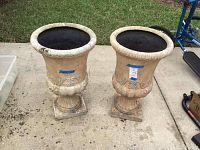 Front view of two Grecian style pedestal planters showing decorative carvings and square bases, resting on concrete ground outdoors.