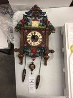 Front view of clock showing colorful resin body, roman numeral face, decorative scrollwork and 2 hanging weights and pendulum.