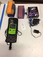 Overall photo showing the green Game Boy Color handheld, three game storage cases, Game Shark video enhancer box with cables, charger, portable speaker, and reading light on a table.