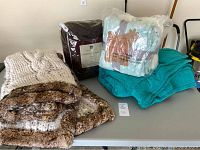 Photo showing four blankets and throws on a table including Pottery Barn chunky knit with faux fur trim, and three Berkshire Blankets throws packed and unpacked.