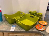 Set of olive green ceramic bakeware including one 4 quart rectangular casserole dish, one 9x9 inch square casserole dish, six small individual servers, and two small pumpkin themed plates arranged on countertop.