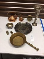 Photo showing all items on a white table: 2 copper kettles, silver-plated platter, tall silver-plated trumpet vase, brass cup, small brass bowl, brass pan with handle, 2 brass shoe-shaped ashtrays.