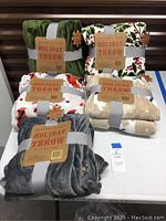 Full set of five Berkshire Holiday Throw fleece blankets arranged on table showing packaging and labels. Two solid color throws (green and gray) and three printed throws with holiday themes: cardinals, polar bears, and holly.