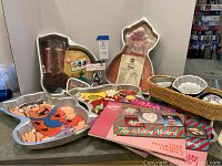 Cake pans displayed on countertop including Power Ranger, Fred Flintstone, Barbie dress, tombstone 'Over the Hill', and cactus shapes with cake decorating books and party favors.