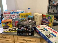 Photo showing eight board games including Super Spirograph, Pictionary, Scrabble, Cranium, Monopoly, The Game of Life Pirates of the Caribbean edition, Clue Haunted Mansion, and a set of markers with coloring books.