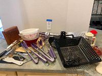 Wide view of the assortment showing bowls, grilling baskets, French press, purple-handled utensils, juicer, and pizza stone.