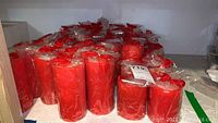 Shelf filled with multiple red pillar candles wrapped in clear plastic, showing floral patterned exterior.
