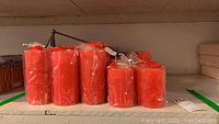 Shelf with multiple red pillar candles by Illuminations, some 6 inch and some 4 inch, all wrapped in plastic packaging, arranged in rows.