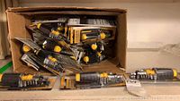 View of multiple ratcheting screwdriver sets, mostly in packaging, with black and yellow handles arranged inside a cardboard box.