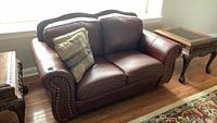 Front view of brown leather loveseat on a hardwood floor under a window, showing rolled arms and nailhead trim