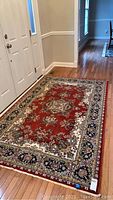 Full view of the rectangular area rug placed on hardwood floor near an entry door, showing red field with intricate floral border in navy and cream tones.