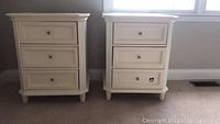 Front view of two white night stands showing three drawers each and metal knob hardware.
