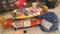 Photo showing yellow and blue Little Tykes picnic bench with red seat, assorted crafting materials and books on and around table, two Winnie the Pooh plush chairs, and wrapped blue play hut structure on floor.
