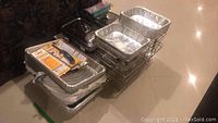 Photo shows stainless steel chafing dishes stacked with aluminum foil pans and metallic racks on floor.