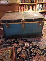 Front view of the vintage metal chest showing blue sides, black top, metal latch and central lock, on the wooden rolling base.