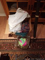 Box with various folded fabrics including floral, teal, and white inside a plastic bag, sitting on patterned rug and wooden floor.