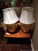 Pair of cream colored ceramic table lamps with white shades on wooden table, cords visible, unplugged.