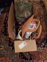 Overview of large cardboard box containing multiple bags and yarn supplies for knitting and crochet crafts placed on patterned carpet