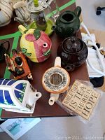 The tea pots arranged on a table showing various styles and colors including a clear glass pot with infuser, green, brown, colorful fruit design, stove-shaped pot, along with a wooden sign.