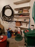 Garage corner with several shelves showing garden hoses, boxes, pruning saws, gardening supplies and green trash can.