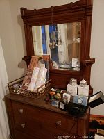 Wide view of vintage dresser with attached wooden mirror, showing paperweight collection, drawer liners in basket, boxed Spode collectibles, and various clocks on dresser top.