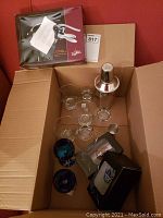 Overview of lot contents in cardboard box showing cocktail shaker, glassware, and corkscrew box.