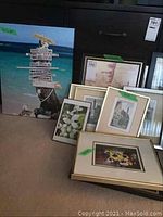 Photo of multiple framed prints leaning against a wall including a large beach directional signpost print and several smaller floral and scenic prints.