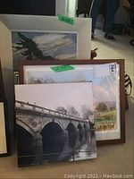 Photo showing three framed artworks including a black and white bridge print, a countryside watercolor, and a larger pine tree painting in a white frame.