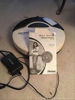 Top view showing Roomba unit, owner’s manual placed in front, and charging adapter at side