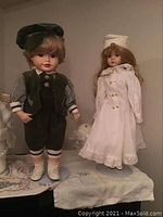 Two porcelain dolls displayed side by side, boy doll in green corduroy with a cap and girl doll in white dress with bonnet, standing on a shelf.