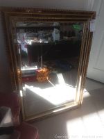 Full view of rectangular gold framed wall mirror leaning against wall, showing its intricate patterned frame and reflective glass surface.
