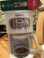White Cuisinart 12 cup coffee maker with digital clock and buttons on countertop, partially shown Melitta coffee box behind it.
